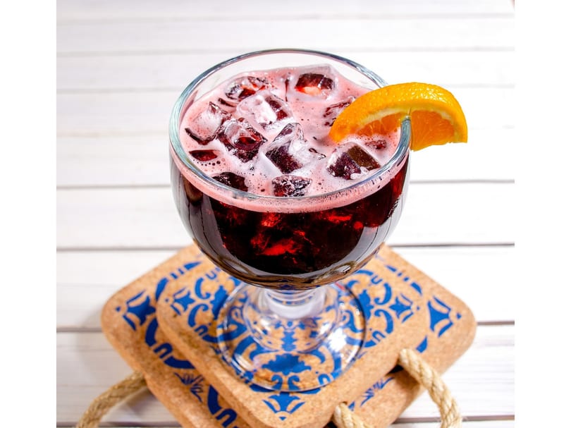 Red sangria with ice and an orange slice, on cork coasters with blue designs.