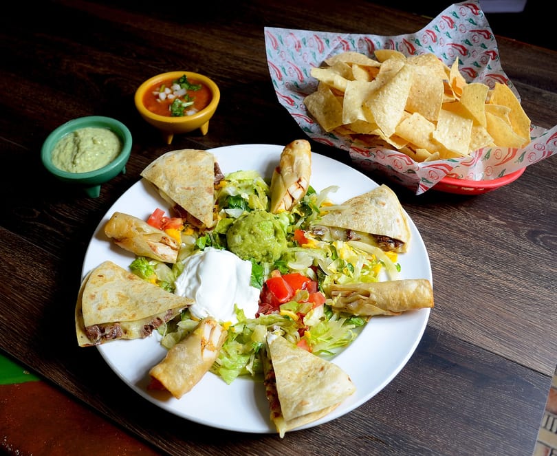 A platter of quesadillas, taquitos, guacamole, sour cream, and salsa, with a basket of chips.