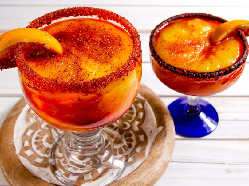 Two vibrant orange and red frozen drinks with chili-lime rims and orange slices on a white wooden table.