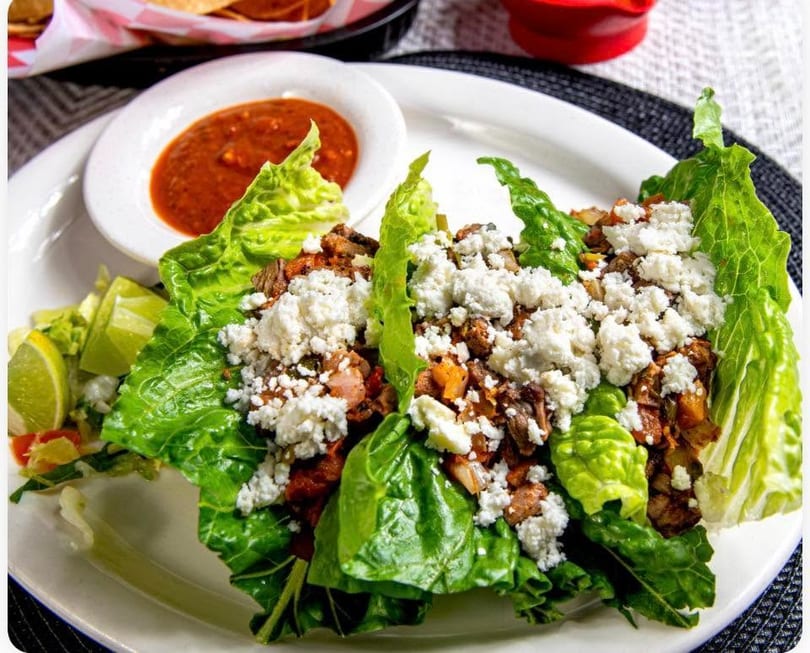 Lettuce wraps filled with meat and crumbled white cheese, served with lime wedges and salsa.