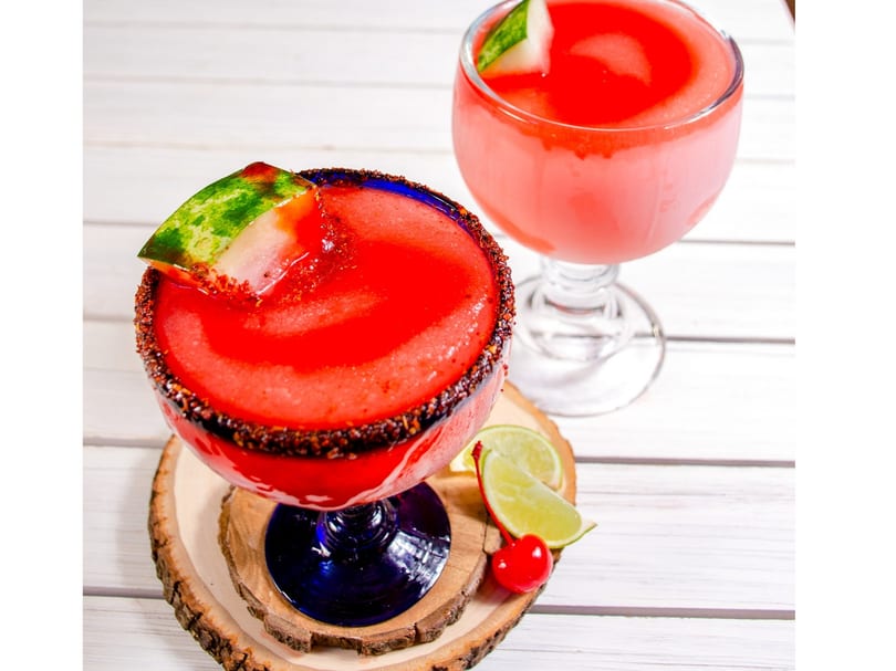 Two red frozen drinks, one garnished with watermelon, lime, and a cherry on a wooden coaster.