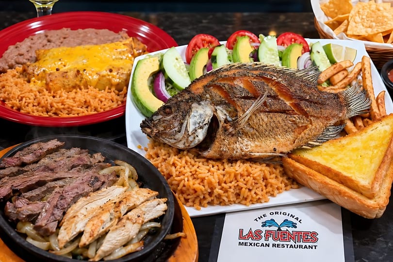 A table laden with various Mexican dishes, including whole fried fish, fajitas, enchiladas, and rice.