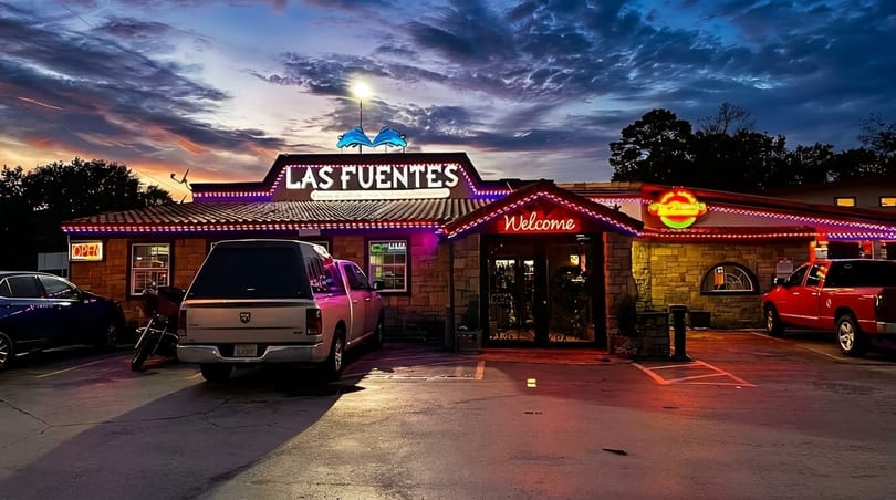 Las Fuentes restaurant exterior at dusk, illuminated by neon lights, with cars parked in front.