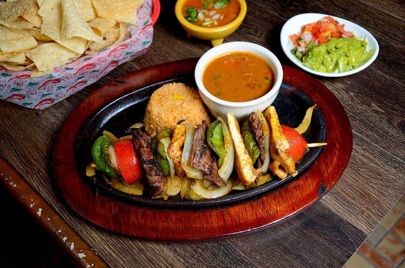 Sizzling fajita skewers with steak, chicken, and vegetables, served with rice, beans, chips, and guacamole.