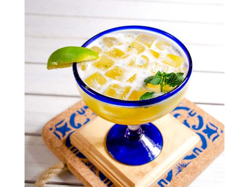 A vibrant yellow drink with ice, lime, and mint in a blue-rimmed glass on a patterned coaster.